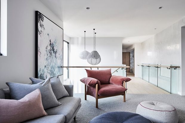 Bright contemporary open-plan living area with a grey sectional sofa, blush and grey cushions, a sculptural burgundy lounge chair, and rounded ottomans. Three woven pendant lights hang above a dining or circulation area, while a glass balustrade overlooks a lower level. A large abstract artwork and pale timber flooring add visual warmth to the refined, light-filled interior.