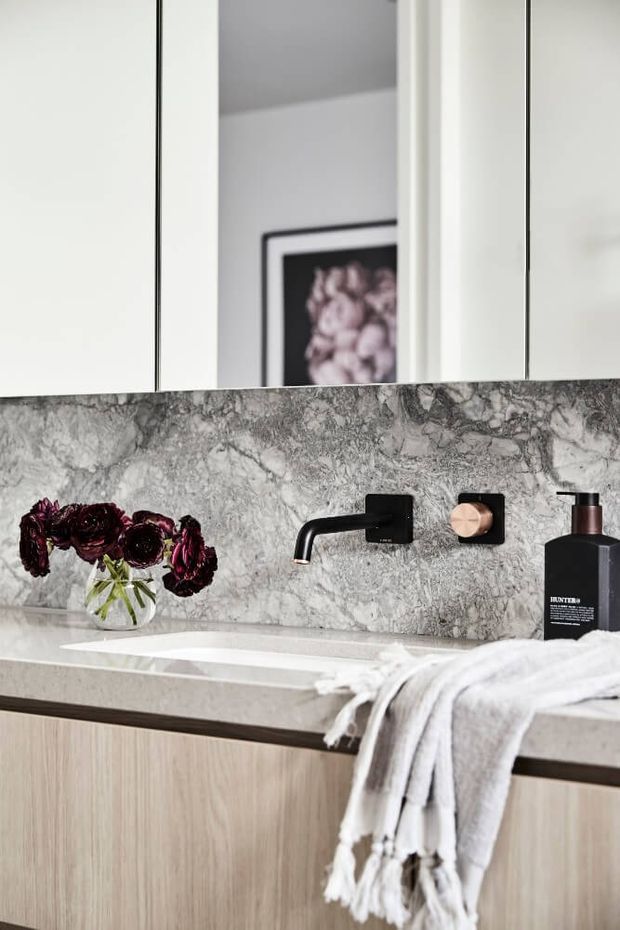 Close-up view of a refined bathroom vanity featuring a pale stone countertop and integrated white basin, light timber cabinetry, a dramatic grey veined stone splashback, matte black wall-mounted tapware, and a large mirror. A folded white towel and dark floral arrangement add contrast.