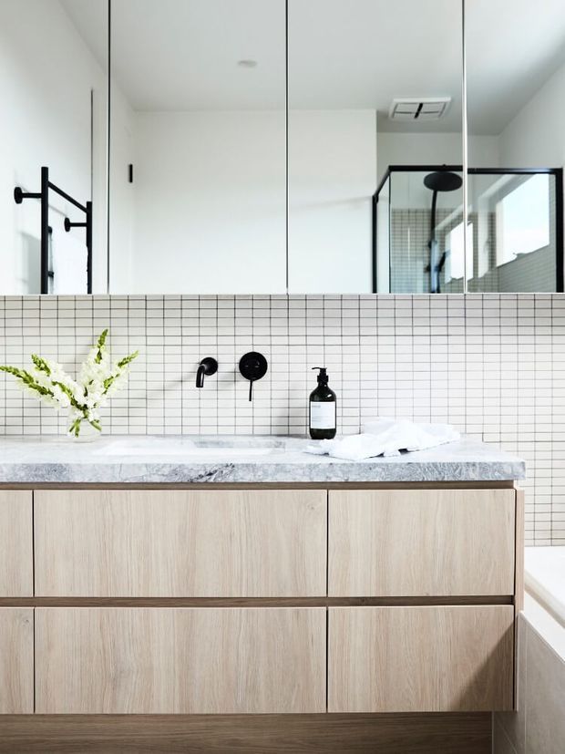 Contemporary bathroom vanity featuring a pale timber floating cabinet with wide drawers, a light grey veined stone benchtop, fine white grid-tile splashback, matte black wall-mounted tapware, and a large mirrored cabinet above. A black-framed glass shower is reflected in the mirror, and a freestanding bath edge is visible at right.