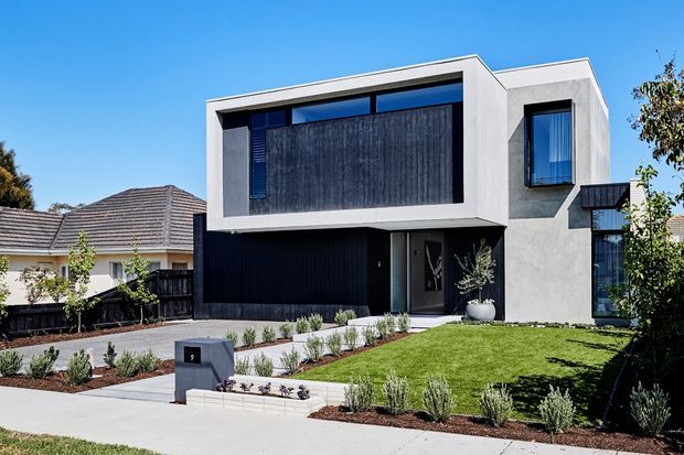 Contemporary two-storey residence with a crisp white rendered facade, deep charcoal-black vertical cladding, large dark-framed windows, and a flat-roofed geometric form. The home is presented from the street with a landscaped front garden, paved driveway, low planting beds, and neighbouring suburban houses visible around it.