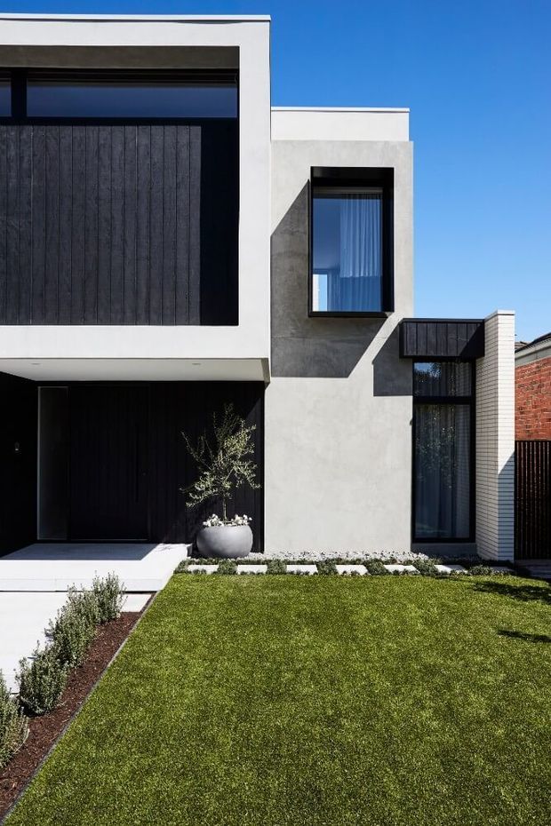 Street-facing view of a contemporary multi-storey townhouse facade with crisp white rendered walls, dark vertical timber-look garage and entry doors, charcoal window frames, projecting box forms, and a neatly landscaped front lawn with low planting and a potted tree. A neighbouring brick wall is visible at the right edge.