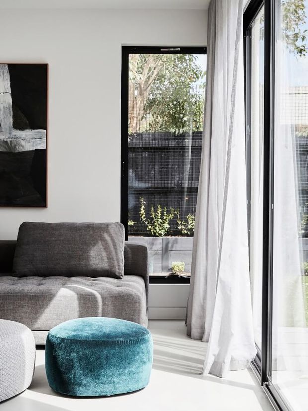 Bright contemporary living room with a grey upholstered sofa, teal round ottoman, pale flooring, white walls, abstract artwork, and full-height black-framed glazing softened by sheer white curtains. Strong natural daylight enters from the large windows and sliding glass door, with a small planted outdoor area visible beyond.