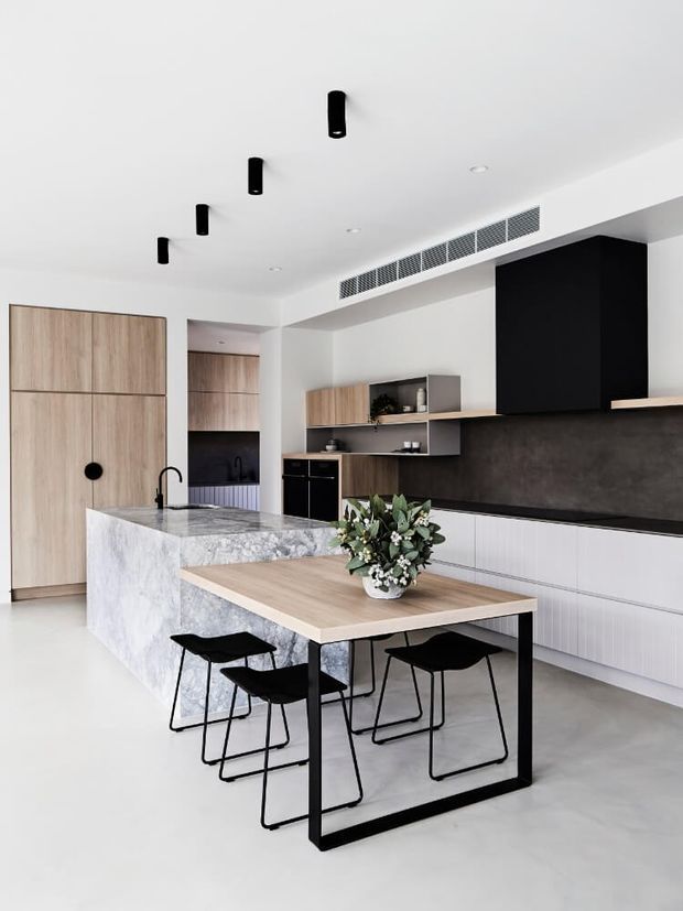 A bright, contemporary kitchen with a large veined stone island, integrated timber cabinetry, dark feature cabinetry and splashback, and a timber dining table with black metal-framed chairs. Minimal black pendant-style ceiling lights and concealed ventilation reinforce the clean, refined design.