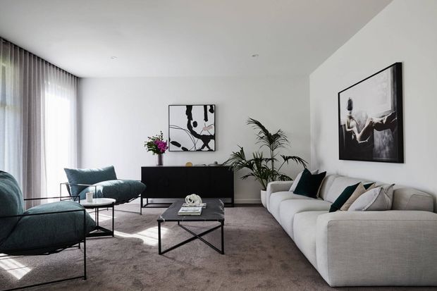 A bright contemporary living room with a large light-grey sectional sofa, dark teal lounge chairs and cushions, a black low-profile media console, glass-topped coffee table, indoor plants, floor-to-ceiling sheer curtains, and monochrome wall art. The space has a soft neutral palette with dark accents and a refined, uncluttered arrangement.