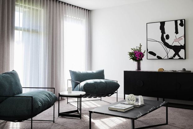A bright, refined living room with floor-to-ceiling sheer curtains, a low black sideboard, teal upholstered lounge chairs, and a minimalist black coffee table. Abstract monochrome artwork and a small floral arrangement provide restrained decorative accents, while strong daylight creates an airy atmosphere.