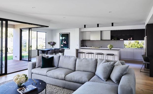 Bright open-plan contemporary living area with a large light-grey sectional sofa in the foreground, opening onto a dining area and a sleek kitchen with a central island and bar seating. Full-height black-framed glazing connects the room to a timber deck and garden, while pale timber flooring, white walls, and dark kitchen cabinetry create a clean, refined interior.