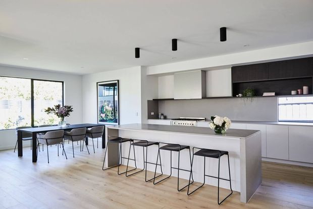 Bright open-plan kitchen and dining area with a large pale island, integrated cabinetry, black linear pendant lights, black-framed stools, and a dining table beside expansive glazing. The room has light timber flooring, white walls, dark grey cabinetry and a clean contemporary fit-out.