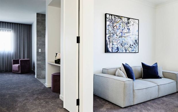 A refined contemporary living room viewed through an open doorway, featuring a large pale upholstered sofa with navy cushions, abstract artwork, white walls, and grey carpet. The adjacent area includes dark curtains, a textured dark feature wall, and a purple armchair, creating a calm, modern sitting area.