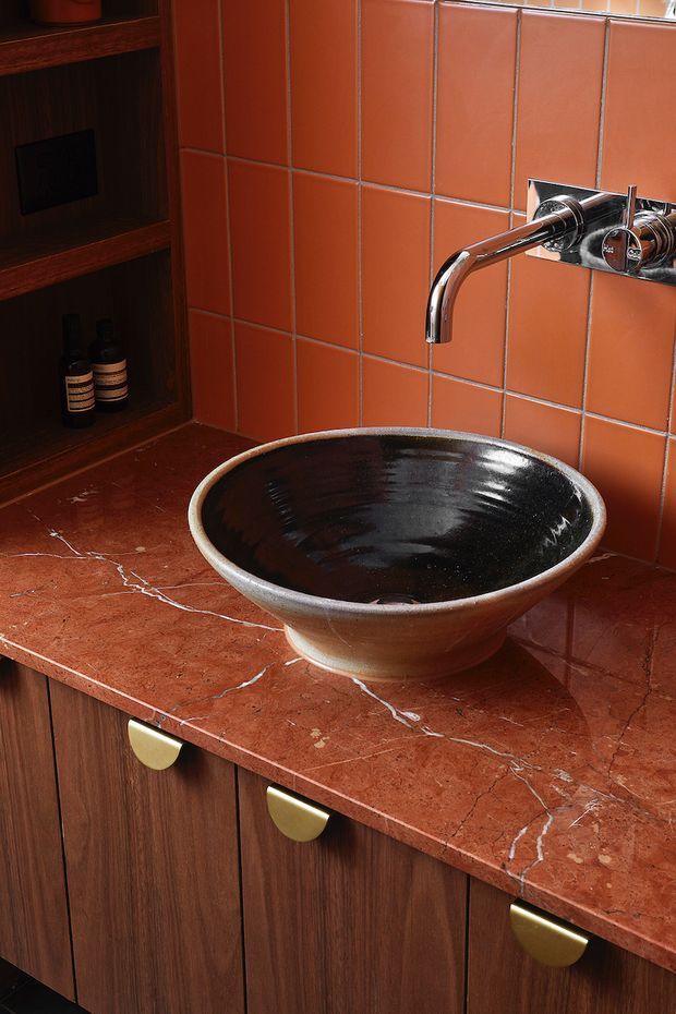 Close-up view of a richly detailed bathroom vanity featuring a vessel basin in dark glazed ceramic, a wall-mounted chrome tap, terracotta-orange tiled splashback, reddish veined stone benchtop, and dark timber cabinetry with brushed brass half-moon pulls. An open timber niche with toiletries is visible at left.