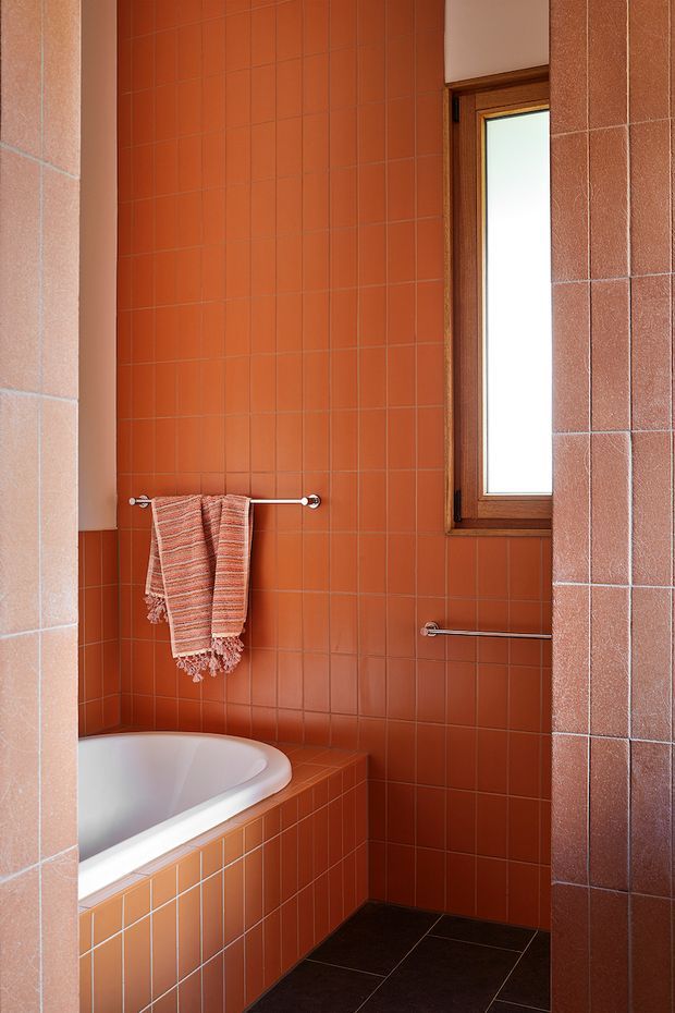 A compact bathroom dominated by terracotta-orange square ceramic wall tiles, with matching tiled bath surround and a dark tiled floor. A white curved bathtub is partially visible at lower left, while a wooden-framed frosted window provides natural light. Chrome towel rails and a woven rust-toned towel add functional and decorative detail.