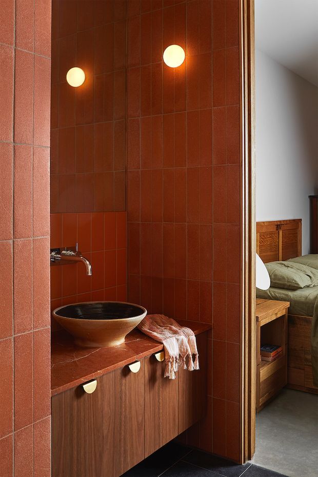 A warm, richly detailed bathroom vanity clad in terracotta-red rectangular tiles, with a dark vessel basin, wall-mounted tap, timber cabinetry, and round wall lights. Through the open doorway, part of a timber-furnished bedroom with a bed is visible.