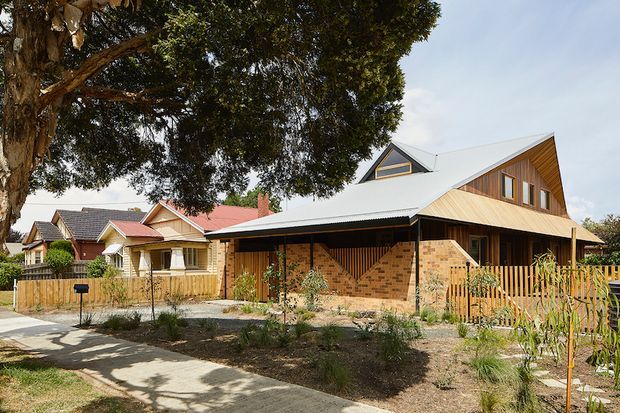 A contemporary residential house viewed from the street, featuring a broad white pitched roof, warm timber-clad upper gable, exposed red-brick walls, dark-framed openings, and a timber picket fence. The home sits within a landscaped suburban streetscape with mature trees and neighbouring traditional houses.