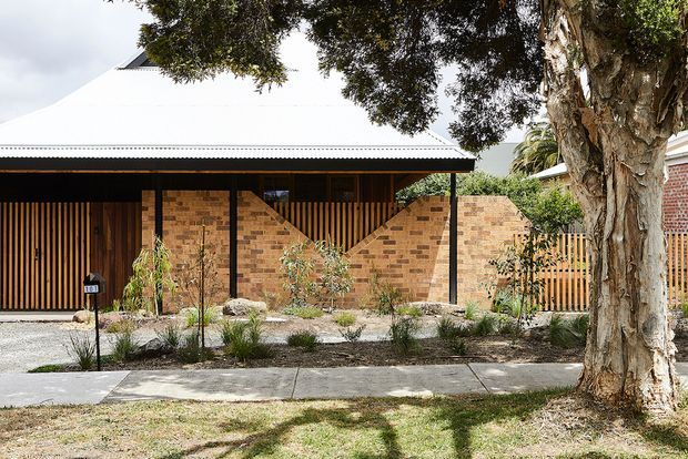 Single-storey residential facade viewed from the street, with a broad white pitched metal roof, dark timber vertical slat screening, warm exposed brick walls, black structural posts, and a landscaped front garden with concrete footpath and mature trees. The image emphasizes the building exterior and its entry screening.