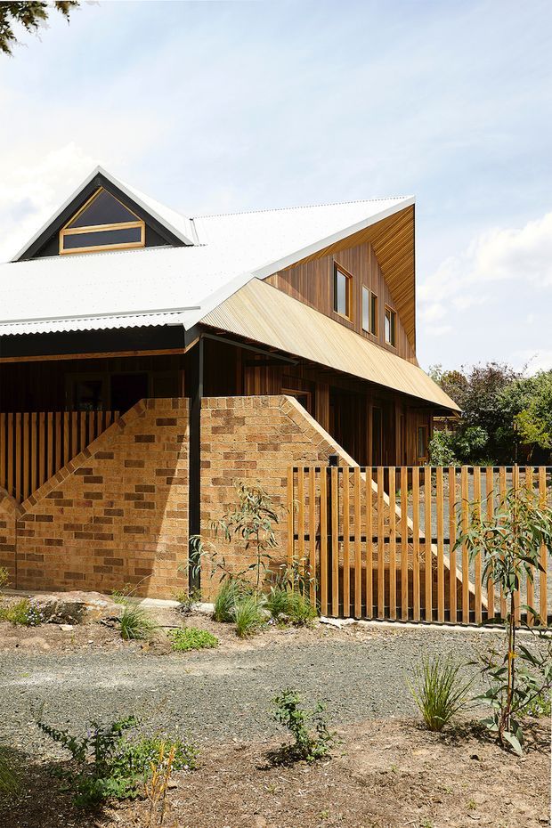 Exterior view of a multi-level timber-clad house with intersecting steeply pitched gabled roofs and a prominent white corrugated metal roof. The facade combines warm vertical timber boards with exposed brick masonry walls, timber-framed windows, and a timber slat fence. A gravel forecourt and sparse landscaped planting occupy the foreground under a bright sky.