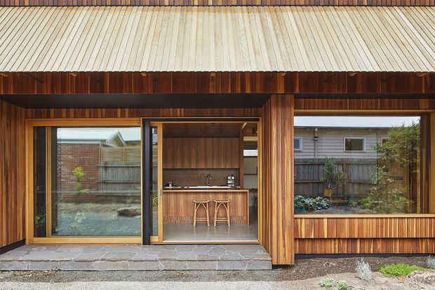 Exterior view of a compact timber-clad pavilion or extension with a corrugated metal skillion roof. Large glazed sliding openings reveal a built-in outdoor kitchen or bar with timber cabinetry, stone benchtop, sink and stools. A covered timber-framed terrace and low planter window create a sheltered connection to the garden.
