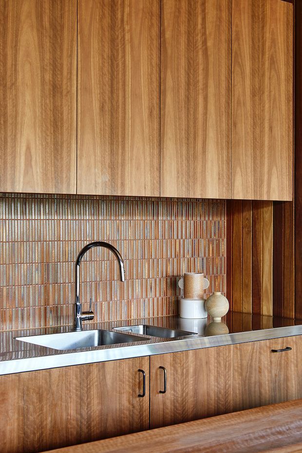 Close-up view of a warm-toned kitchen fitted with extensive natural timber cabinetry, a stainless-steel double sink and gooseneck tap, and a vertically arranged narrow glazed tile splashback in earthy brown and rust tones. The cabinetry has simple flat fronts with dark metal handles, while timber wall panelling continues at the side. A timber benchtop or table edge is visible in the foreground.