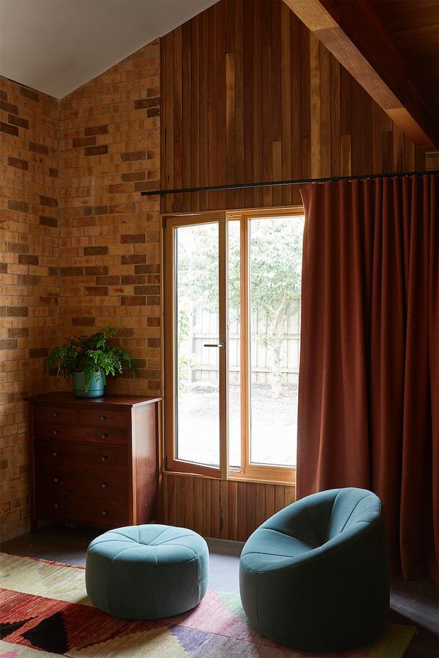 A warm, intimate sitting area with exposed red brick walls, dark vertical timber wall panelling and a steep timber-lined ceiling. A tall timber-framed glazed door/window brings in natural light, partly covered by a deep rust curtain. Two rounded blue-green upholstered lounge chairs and a matching ottoman sit on a colourful patterned rug beside a dark timber chest of drawers and potted plant.