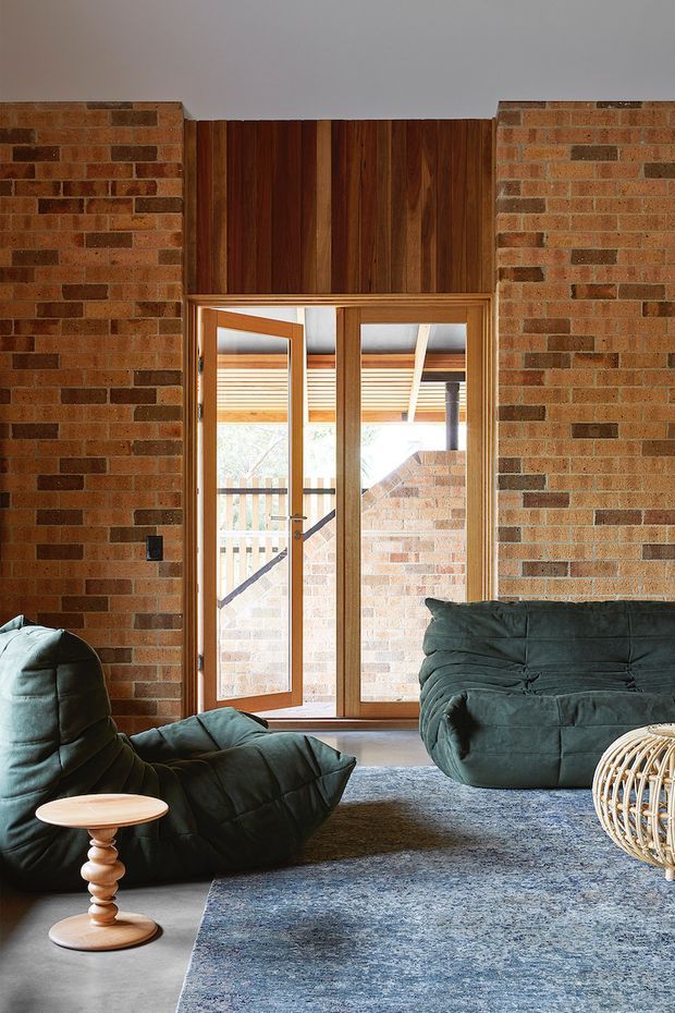 A warm, brick-lined living room with a timber-framed glazed door opening to an outdoor area. Dark green low-profile upholstered lounge seating surrounds a small sculptural timber side table, with a large blue-grey textured rug over a smooth grey floor. Exposed brick walls and a rich vertical timber feature panel create a tactile, mid-century-inspired interior.