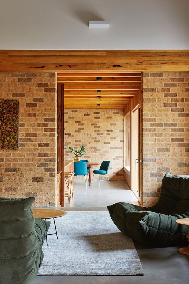 A warm, mid-century-inspired living room with exposed brick walls, timber ceiling beams and lining, and a wide opening into a bright adjoining dining or workspace area. Deep green upholstered lounge chairs, a low round timber coffee table, patterned wall art, and a pale area rug create a relaxed, richly textured interior. The adjoining space repeats the brick and timber finishes and contains a small table with blue and dark teal chairs.