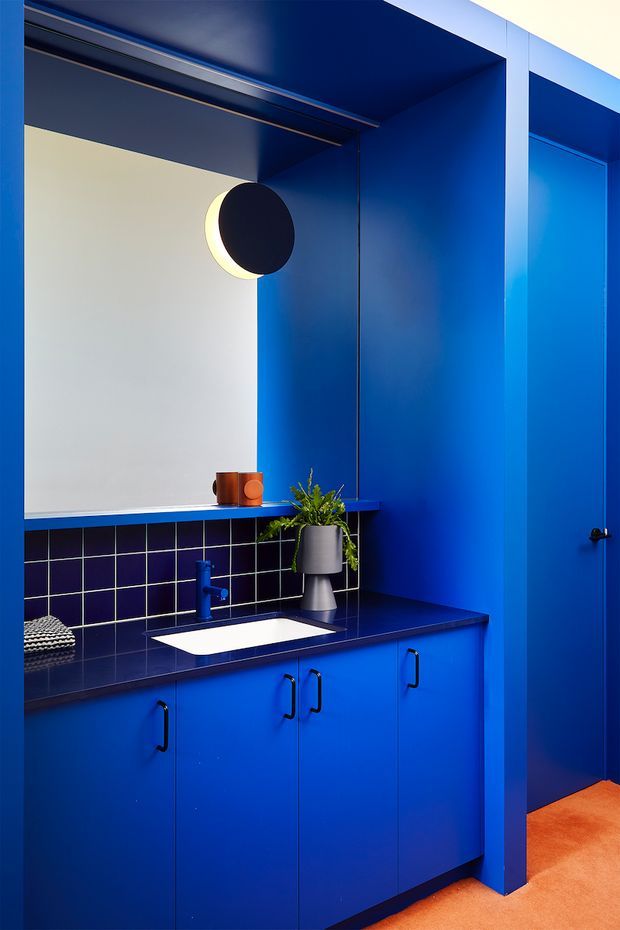A compact, vividly blue bathroom vanity area with floor-to-ceiling blue cabinetry and wall surfaces, a dark blue tiled splashback, white integrated basin, blue tapware, round wall light, and a small potted plant. An orange floor covering provides a strong contrasting accent, while a blue door is visible at right.