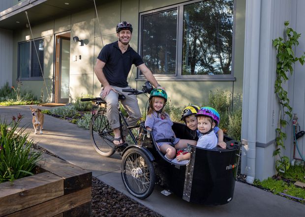 A family with a parent riding a cargo bicycle and three children seated in its front carrier outside a contemporary home. The scene shows the home's modern facade, large windows, covered entry, concrete path, planted garden beds, and a grey exterior wall with climbing greenery.