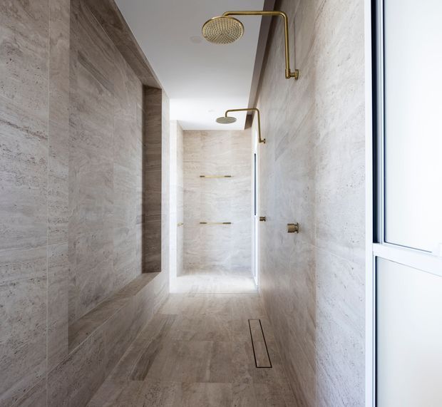 A bright, narrow walk-in shower room clad in continuous pale beige stone-look tile across the walls and floor. Two shower positions with brushed brass/gold rain showerheads and controls are visible, along with recessed wall niches, linear floor drains, and a frameless glass partition. A white-framed window or door provides natural light at the right.