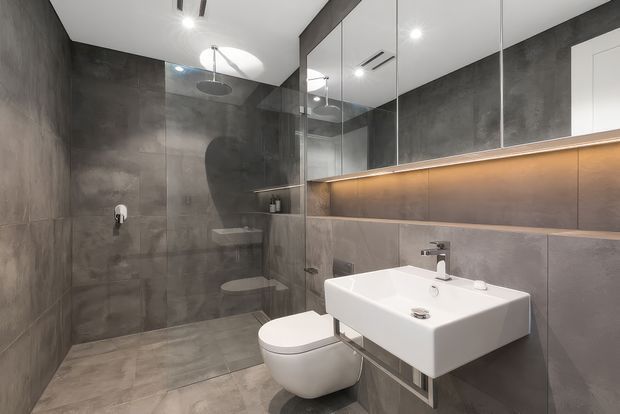 Contemporary bathroom with full-height dark grey stone-look wall and floor tiles, a frameless glass walk-in shower, wall-hung toilet, and floating white rectangular vanity basin. Mirrored wall cabinetry and a warm illuminated recessed shelf add depth and contrast to the minimalist space.