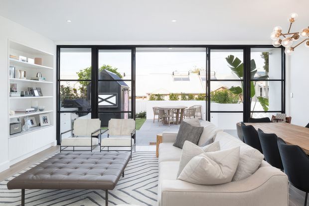 Bright, spacious open-plan living and dining room with full-height black-framed glazing opening to an outdoor entertaining area. The room features a large cream sectional sofa, tufted leather ottoman, contemporary lounge chairs, a timber dining table with dark dining chairs, built-in white display shelving, a geometric rug, and a sculptural brass-and-glass pendant light. The outlook includes a landscaped courtyard and neighbouring residential buildings.