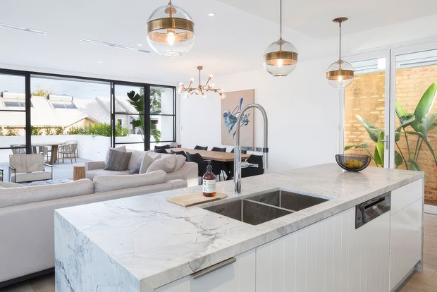 Bright open-plan kitchen and living area with a large white veined stone island, integrated double sink and dishwasher, white vertical-panel cabinetry, sculptural brass-toned pendant lights, and a neutral sectional sofa. Black-framed glazing opens the room toward a small outdoor terrace, with a dining area and contemporary decorative lighting beyond.