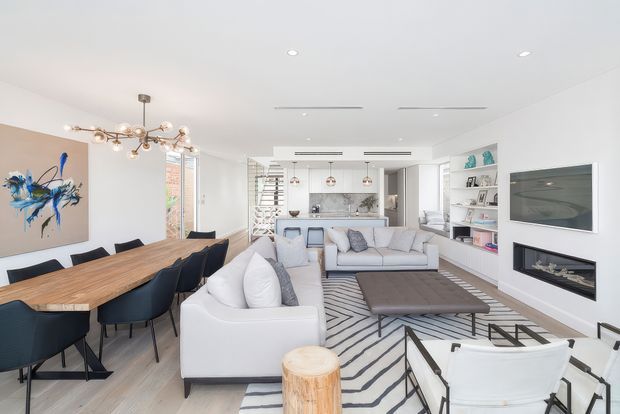 Bright, open-plan contemporary living and dining area with a white sofa arrangement, black-and-white geometric rug, timber dining table, dark upholstered dining chairs, built-in shelving, and a wall-mounted television above a recessed fireplace. A compact kitchen is visible beyond the living room, with large glazed openings and abundant natural light.