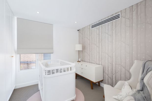 Bright, softly styled nursery featuring a white crib, white mid-century-inspired dresser, upholstered rocking chair, pale patterned feature wallpaper, and a large roller blind. The room has light grey carpet, white walls and ceiling, recessed lighting, and a calm neutral palette with subtle pink accents.