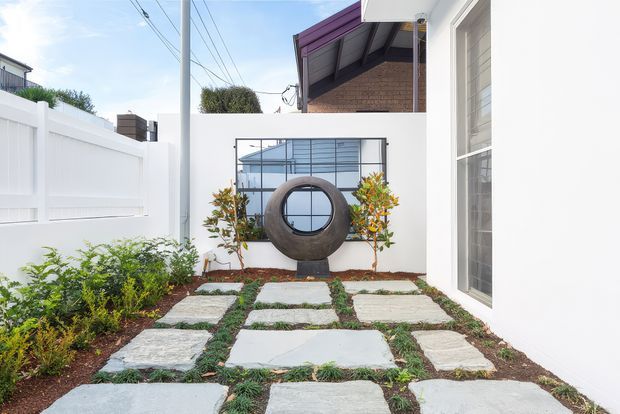 A compact landscaped courtyard or side garden beside a contemporary white building. Large irregular pale stone pavers are set into low green groundcover, bordered by planted beds and a white privacy fence. A circular sculptural feature sits against the rear wall beneath a horizontal metal-framed window, with a tall glazed opening on the right. Overhead utility wires and neighbouring rooflines are visible beyond the enclosure.