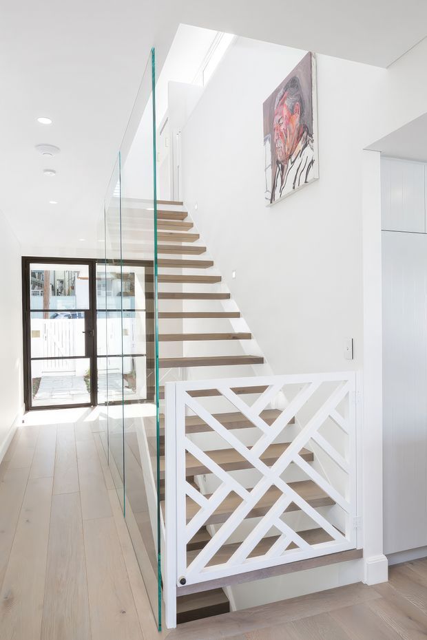 Bright contemporary residential stairway and landing with floating light timber stairs, clear glass balustrade, and a distinctive white geometric-patterned stair guard. White walls and ceiling, pale timber flooring, recessed ceiling lights, a dark-framed glazed entry door, and a colourful artwork create an airy, minimalist interior.