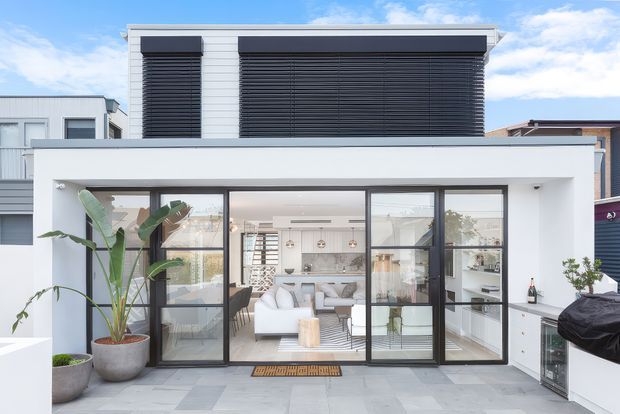 Exterior view of a contemporary two-storey residence with a crisp white rendered facade, extensive black-framed glazing, and dark horizontal exterior blinds. The ground floor opens onto a paved outdoor terrace and reveals a bright open-plan living and kitchen area, while potted tropical plants and built-in outdoor cabinetry frame the foreground.