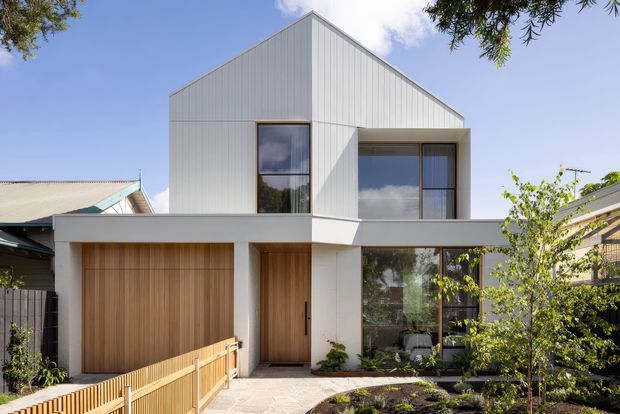 Street-facing view of a compact contemporary two-storey residence with a prominent gabled roof form, white vertical metal cladding, timber garage and entry doors, broad glazing, and a small landscaped front garden. The house is integrated into a suburban streetscape with neighbouring homes visible on both sides.