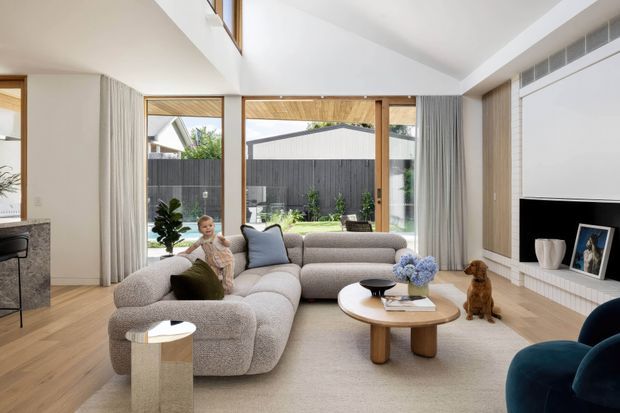 Bright contemporary living room with a large rounded sectional sofa, timber flooring, floor-to-ceiling glazing, sheer curtains, a sculptural timber coffee table, and a built-in fireplace wall with timber detailing. The room opens to a private courtyard and is finished in soft neutral tones with warm timber accents.