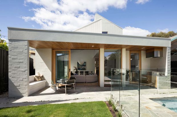 Contemporary single-storey rear extension with a broad flat-roofed outdoor living canopy, white painted masonry walls, warm timber soffits and posts, extensive glazing, and a glass pool safety balustrade. A covered patio with outdoor seating opens directly to a lawn and swimming pool, while a living room is visible through the glazed doors. A pitched gable roof rises behind the low extension.