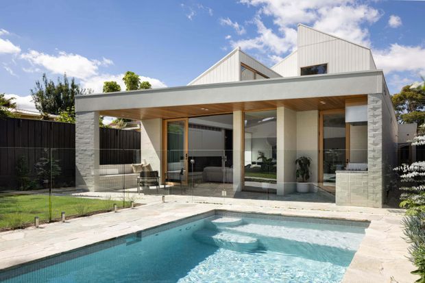 Modern low-rise residence viewed from the backyard, featuring a flat-roofed covered outdoor terrace with timber soffit, extensive glazing, pale masonry walls, and a swimming pool in the foreground. The house includes angular pitched roof forms behind the terrace and a landscaped garden setting.