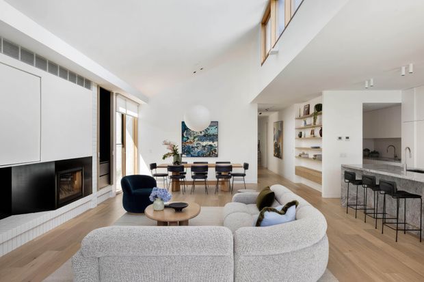 Bright open-plan contemporary living space with a large curved sectional sofa in the foreground, a dining area beyond, and a kitchen with a stone island and bar stools to the right. The room features light timber flooring, white walls, a black fireplace surround with integrated firebox, built-in shelving, extensive glazing, and a partial double-height ceiling with clerestory-style upper windows.