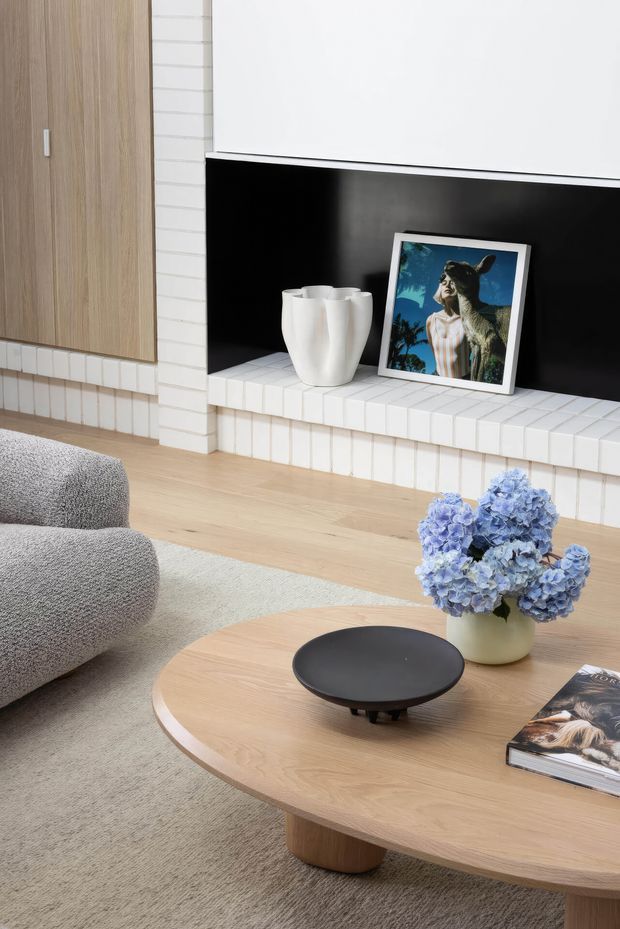 A bright, refined living room vignette featuring a large rounded timber coffee table, a textured curved grey sofa, pale carpet, and a black shallow decorative bowl. The background includes a white tiled fireplace surround and hearth with a glossy black inset, light timber built-in cabinetry, a white sculptural vase, and framed artwork. Blue hydrangeas add colour to the otherwise neutral palette.