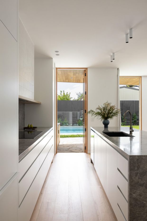 Bright, narrow contemporary kitchen with white full-height cabinetry, integrated appliances, pale timber flooring, and dark grey stone benchtops. A central island with a black undermount sink and curved tap faces a glazed door opening to a pool courtyard. The room has a clean white ceiling with recessed and cylindrical track-style spotlights, timber-framed openings, and a minimal, refined palette.