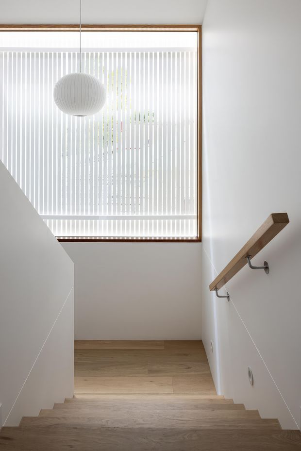 A bright, minimalist stairway with pale timber treads and flooring, white walls, a simple timber handrail on metal brackets, and a large window fitted with vertical white blinds. A round white pendant light hangs in front of the window, creating a calm, airy interior.