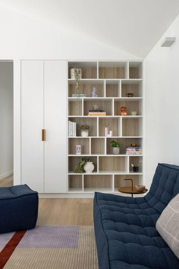 Contemporary living room with a built-in wall unit combining tall white storage cabinetry and open timber-toned shelving. The room features deep navy tufted seating, a small round wood side table, light timber flooring, and layered area rugs. The shelving displays books and decorative objects, while a sloped ceiling and clean white walls create a bright, refined interior.