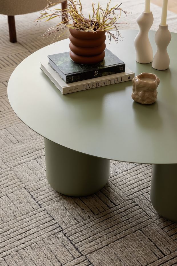 Close-up interior styling vignette centered on a large rounded sage-green coffee table with cylindrical pedestal legs. The table holds stacked design books, a sculptural brown planter with fine foliage, two cream sculptural candleholders, and a small organic ceramic vessel. A textured beige geometric rug and partial upholstered seating surround the table, creating a warm, refined contemporary setting.