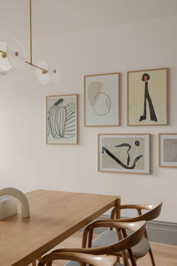A warm, refined dining area with a light timber dining table and sculptural wooden chairs. The soft beige wall features a curated arrangement of abstract framed artworks, complemented by a contemporary brass-and-opal-glass linear pendant light. A grey dado or wainscot detail runs along the lower wall.