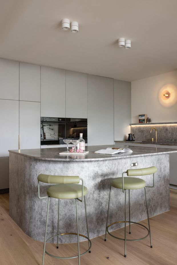 A refined contemporary kitchen with full-height pale cabinetry, integrated black appliances, a large textured grey stone island, and a matching stone splashback. Two pale green upholstered bar stools sit at the island, with warm brass tapware, discreet under-shelf lighting, timber flooring, and sculptural wall lighting creating a polished yet inviting atmosphere.