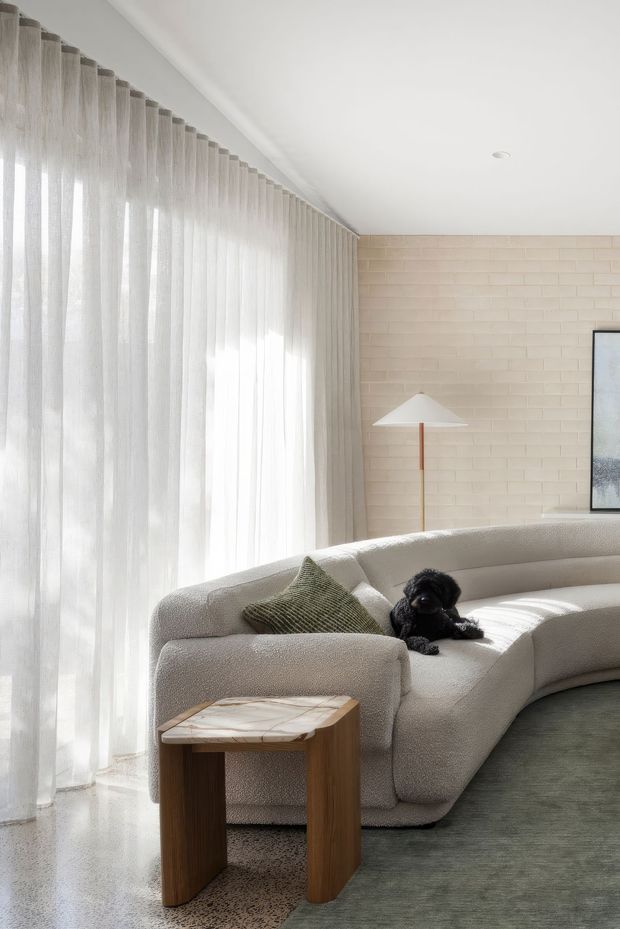 Bright, refined living room with a large curved cream boucle sectional, sheer full-height curtains, a pale exposed brick feature wall, sculptural floor lamp, marble-topped timber side table, and muted green rug. Natural light creates an airy, calm atmosphere.
