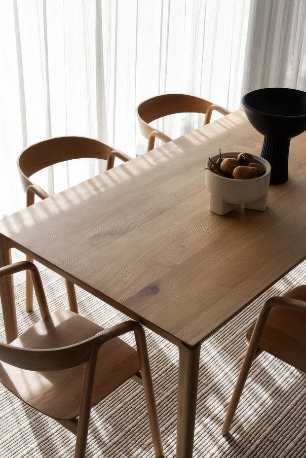 A softly lit dining area with a large light timber dining table surrounded by curved timber dining chairs. Sheer floor-to-ceiling curtains diffuse natural light, while a textured neutral rug, black sculptural bowl, and ceramic fruit vessel add understated decorative accents. The space has a calm, refined, contemporary natural aesthetic.