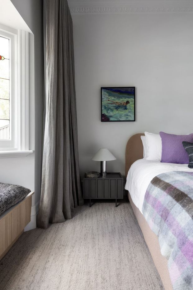 A refined bedroom interior with a white wall and decorative ceiling cornice, tall window with layered grey curtains, grey carpet, upholstered bed with white bedding and purple accent pillows, a dark bedside cabinet, and a small table lamp. A framed artwork hangs above the bed, while a built-in window seat is partially visible at left.