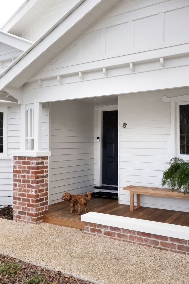 Front entry porch of a white weatherboard home with a prominent gabled roof, exposed painted timber rafters and brackets, red brick columns and low retaining walls, dark front door, timber decking, and a simple built-in timber bench. The image is photographed from outside and focuses on the covered entry and facade.